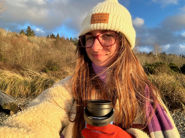 A woman outdoors wearing a cream ribbed knit beanie with a brown 'Black Rifle Coffee Co.' patch, glasses, a cream sherpa jacket, and a purple and pink striped blanket, holding a red insulated tumbler with a black lid.